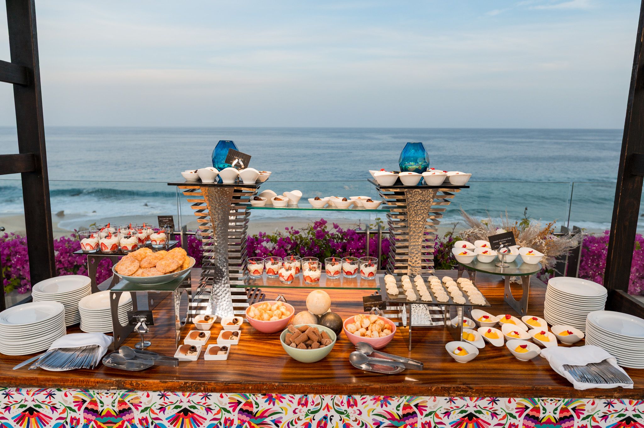 Elegant oceanfront buffet table with desserts, pastries, and floral decor overlooking a scenic beach and blue sea.