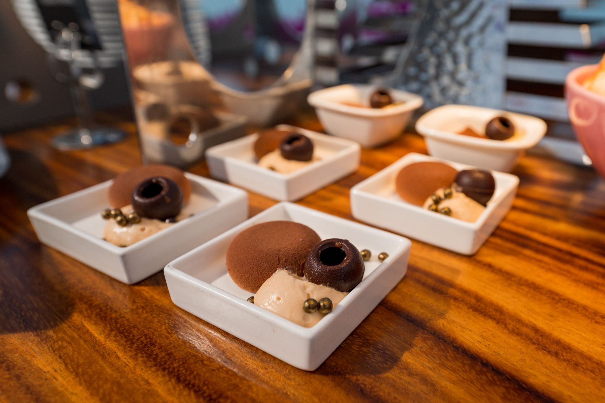 Six white square dishes with chocolate desserts and garnishes displayed on a wooden table.
