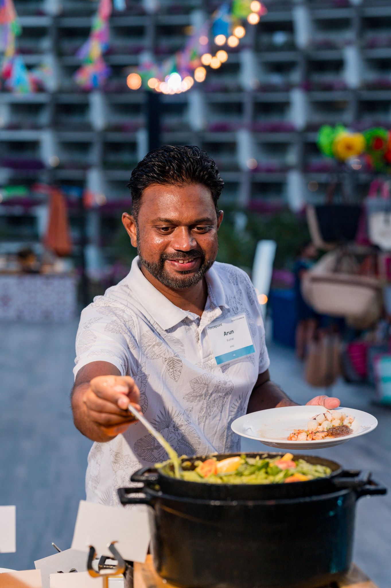 A smiling man in a white polo serves food from a large pot at an outdoor catered event with festive decorations.