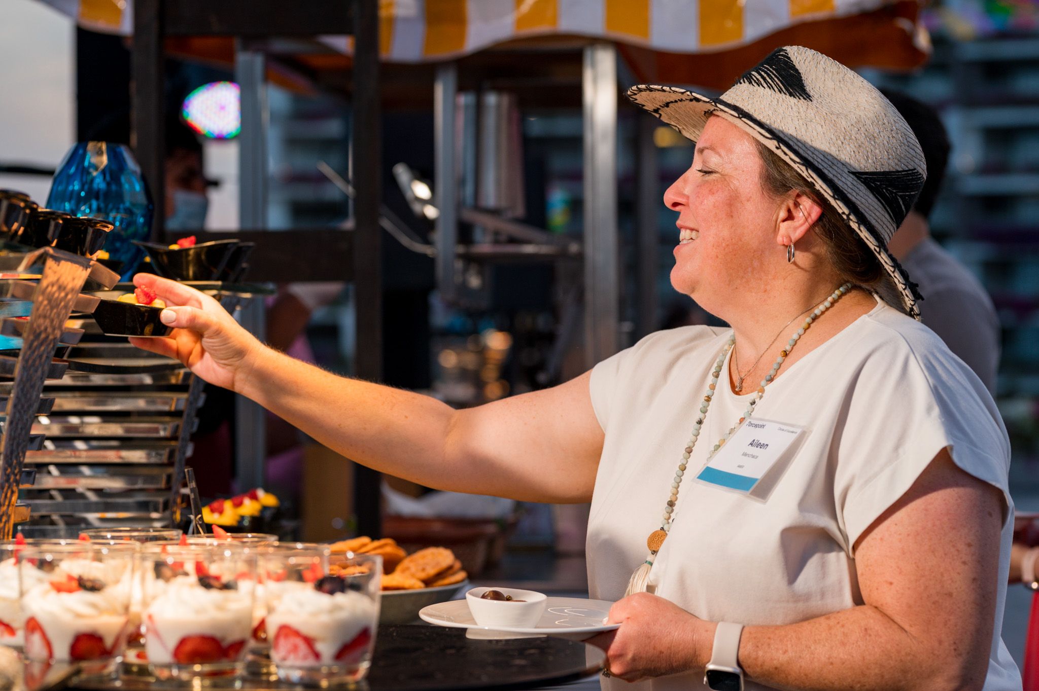 Woman in cowboy hat serving strawberry desserts at a buffet station, smiling while reaching for food display.