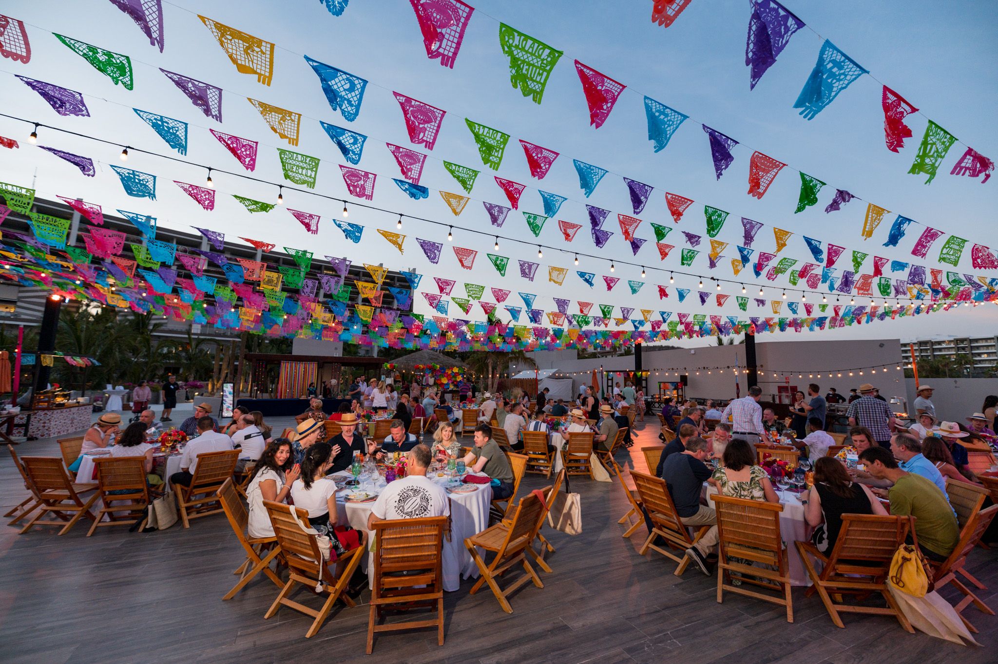Outdoor dining event with colorful papel picado flags overhead, guests seated at wooden tables under a festive, vibrant canopy.