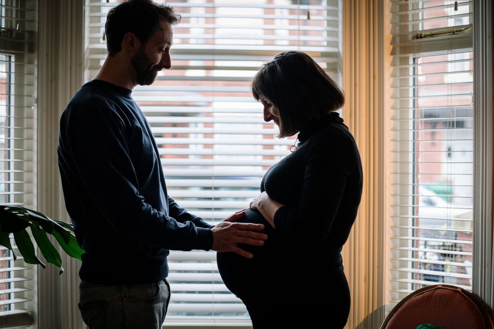 Silhouette of a couple standing by a window, man tenderly touching pregnant woman's baby bump indoors.