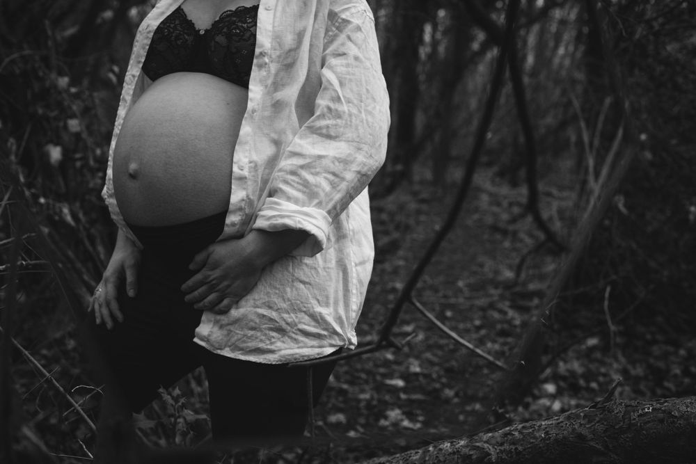 Black and white photo of a pregnant woman outdoors, showing her bare baby bump while holding a pot in the woods.