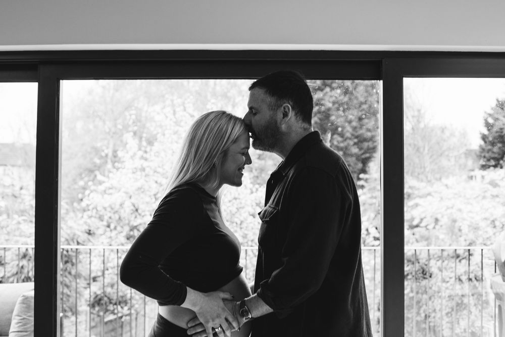 Black and white photo of a pregnant couple sharing a tender moment by a window.