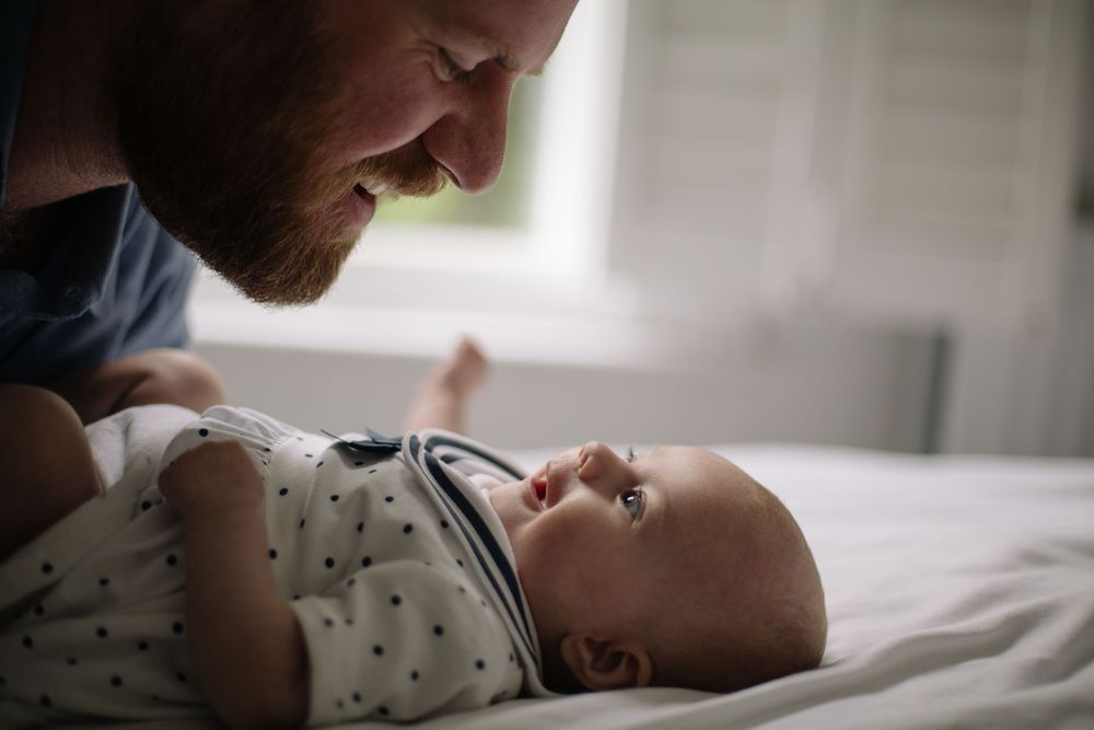 A bearded father smiles lovingly at his newborn baby lying on a white bed, sharing a tender bonding moment.