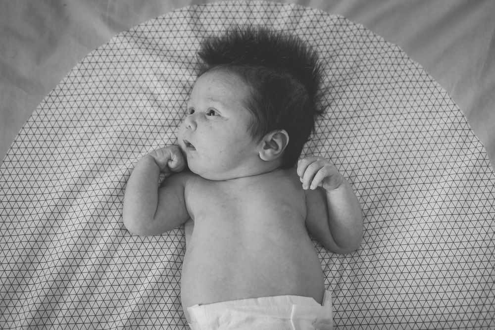 Black and white photo of a newborn baby lying on its back on a patterned blanket, looking alert and curious.