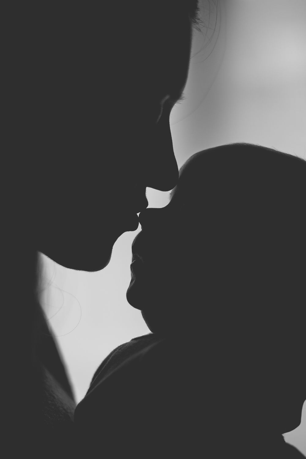 Black and white silhouette of a parent tenderly kissing their newborn baby in an intimate portrait.