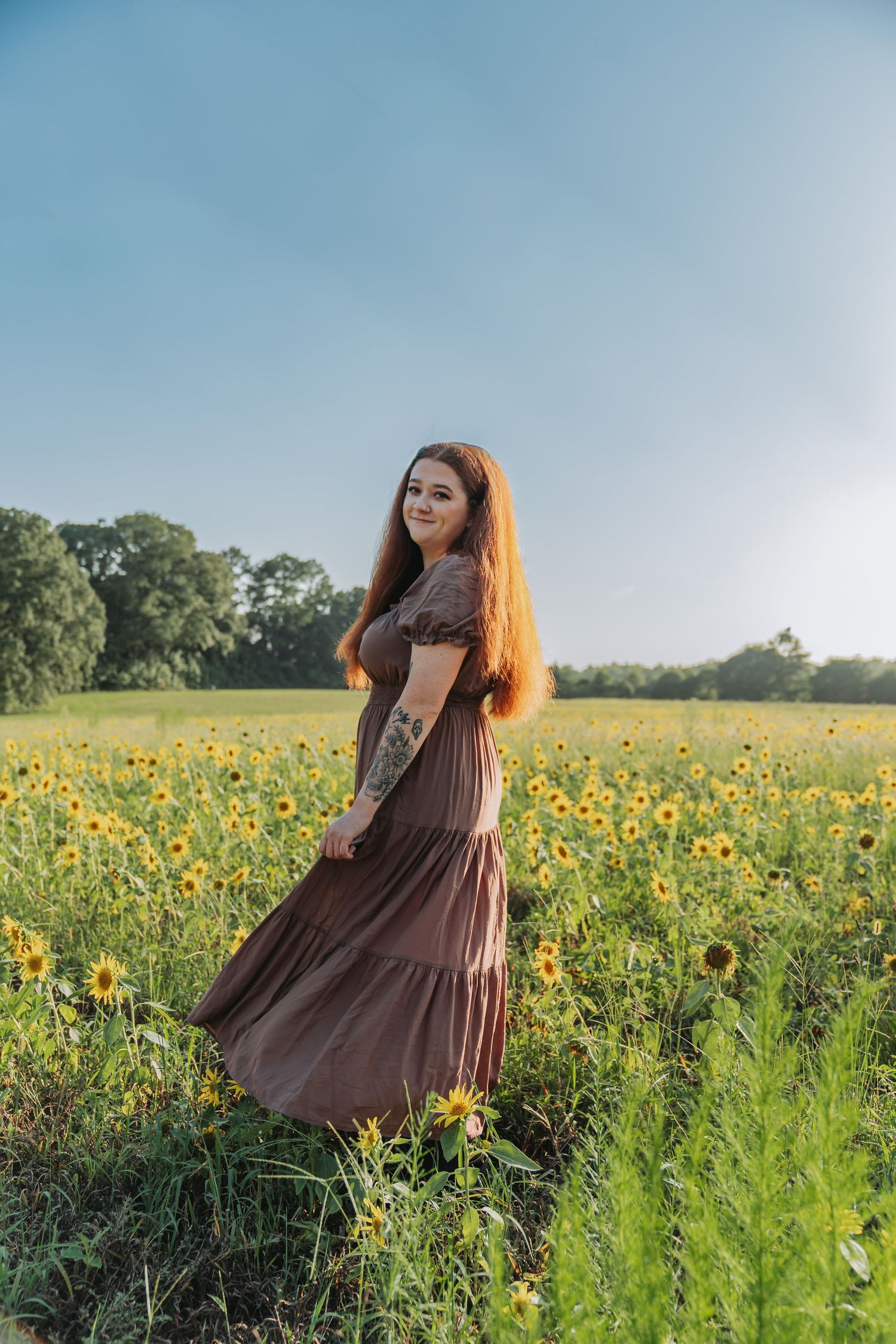 Young woman in brown maxi dress standing in sunflower field, long red hair, tattoo visible, sunny day with blue sky.