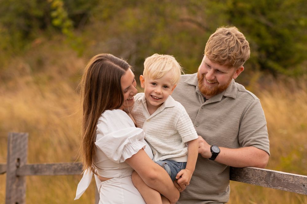 A young family with a toddler boy poses by a wooden fence in a golden autumn field.