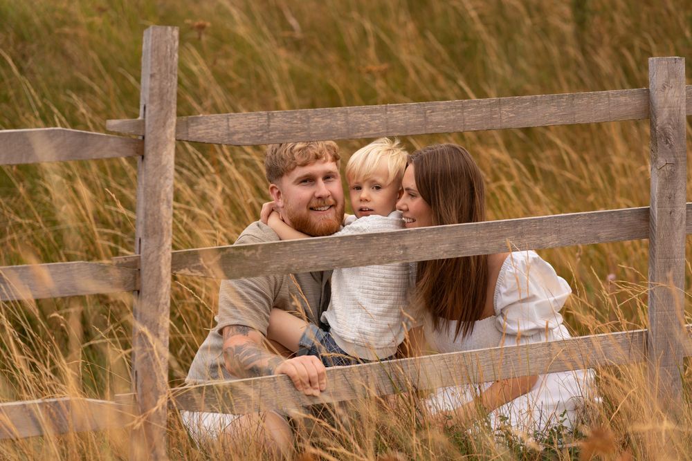 A happy family of three sits by a rustic wooden fence surrounded by golden tall grass fields.