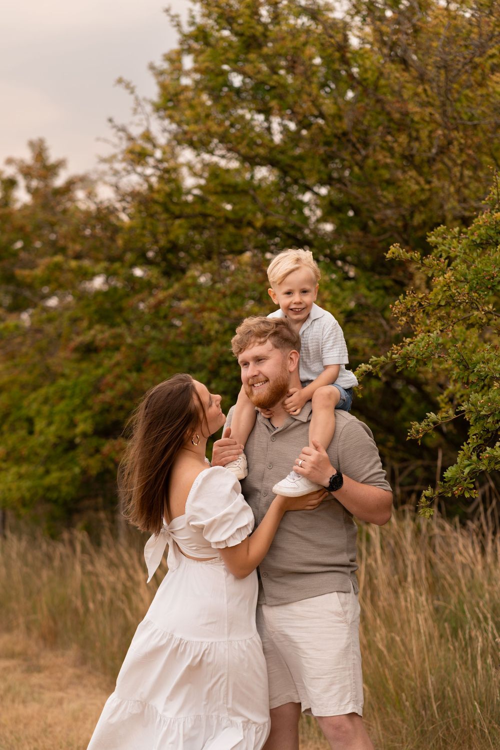 Happy family outdoors, mother in white dress and father carrying toddler son on shoulders in a green field.