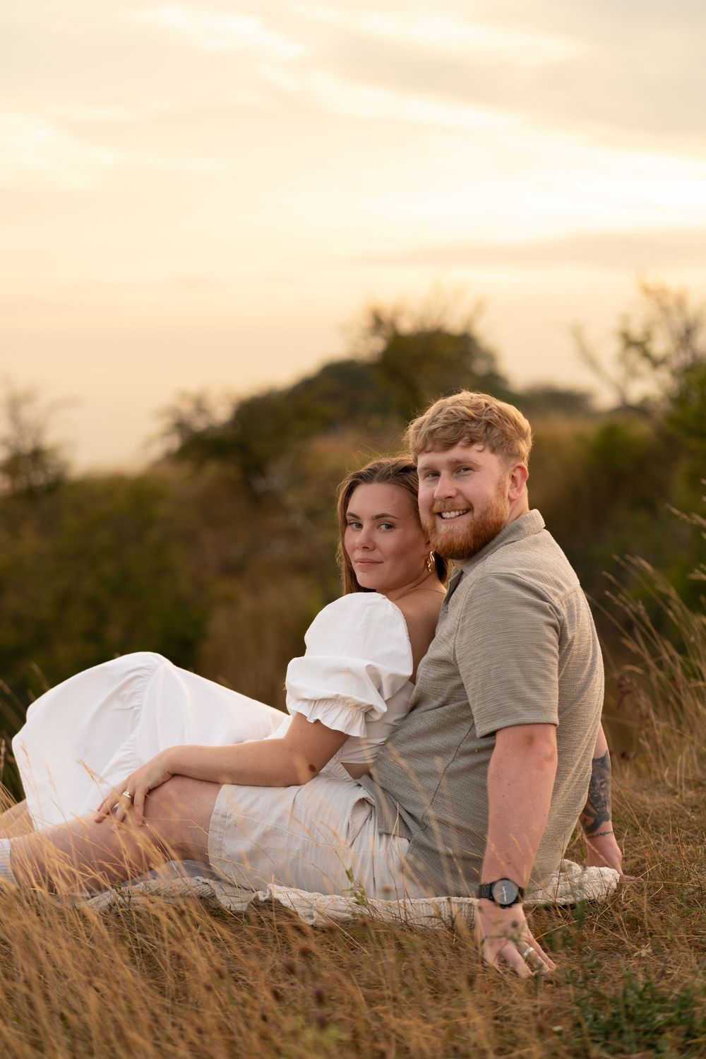 Couple sitting together in golden grass field at sunset, woman in white dress leaning against man in khaki shirt.