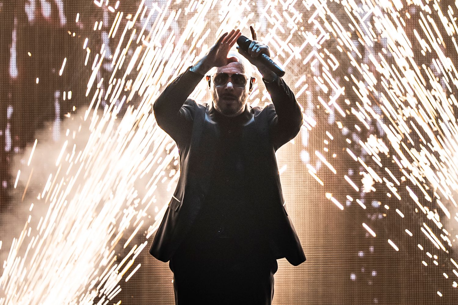 Performer on stage with dramatic pyrotechnic sparks shower, hands raised above head in dark concert setting.