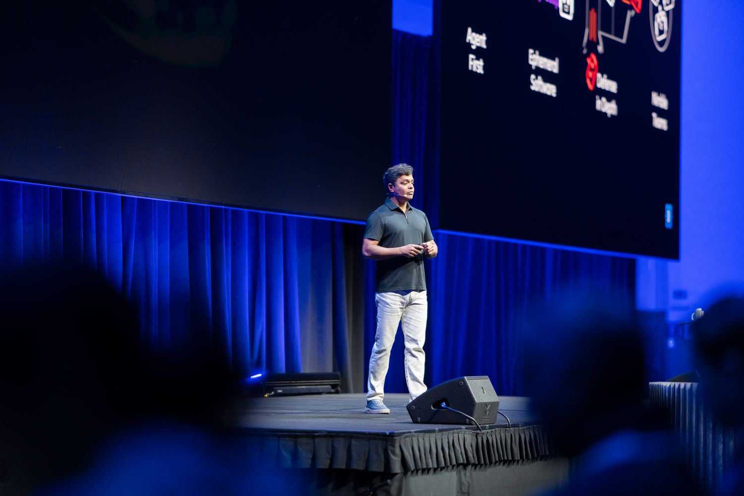 A speaker presents on stage at a conference, gesturing toward a large dark screen displaying a diagram.