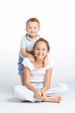 A smiling girl sits cross-legged on white background while her younger brother stands behind her with hands on her shoulders.