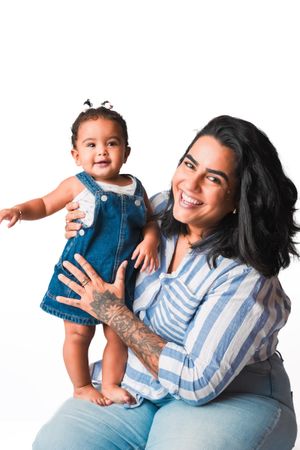 Smiling woman with tattoos holds a happy baby girl in a denim dress against a white background.