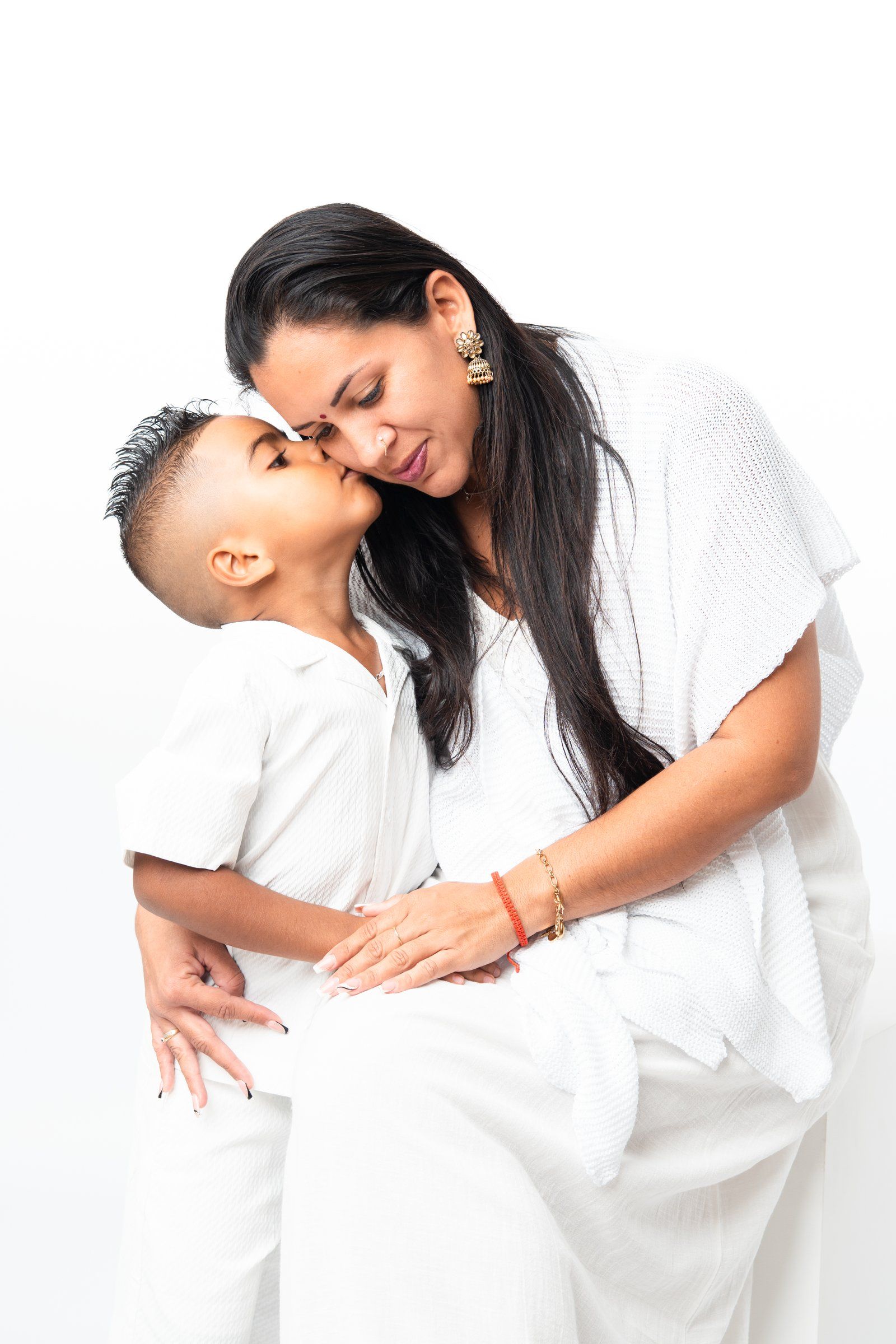 A young boy kisses his mother on the cheek as they share a tender moment, both dressed in white.