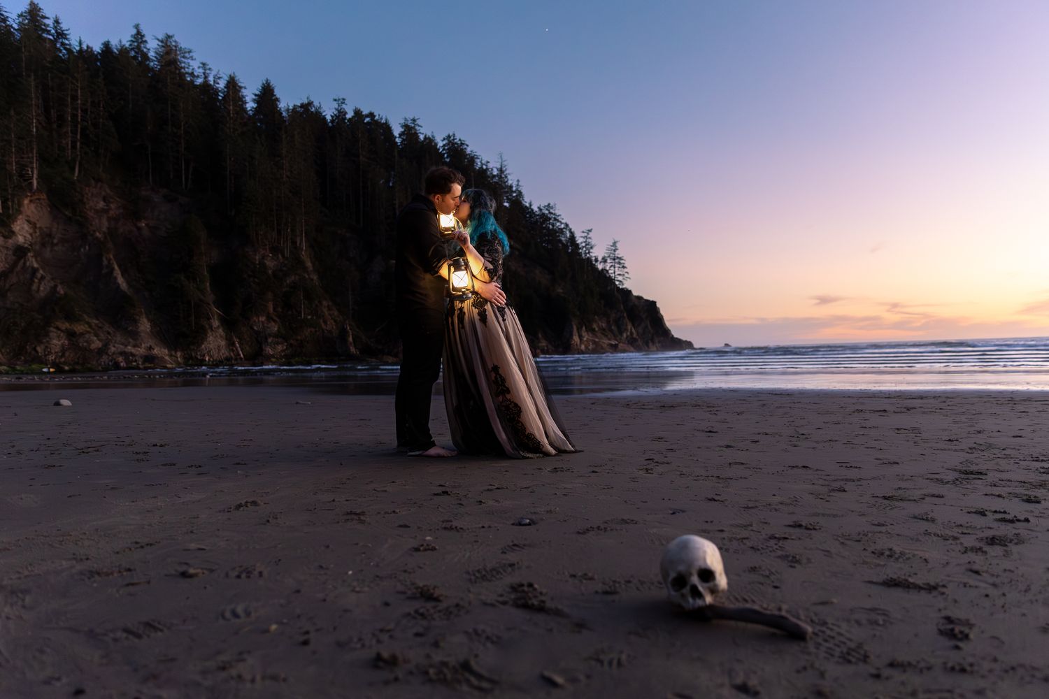 Couple standing on a reflective sandy beach with dramatic cliffs and evergreen trees in the background at sunset.