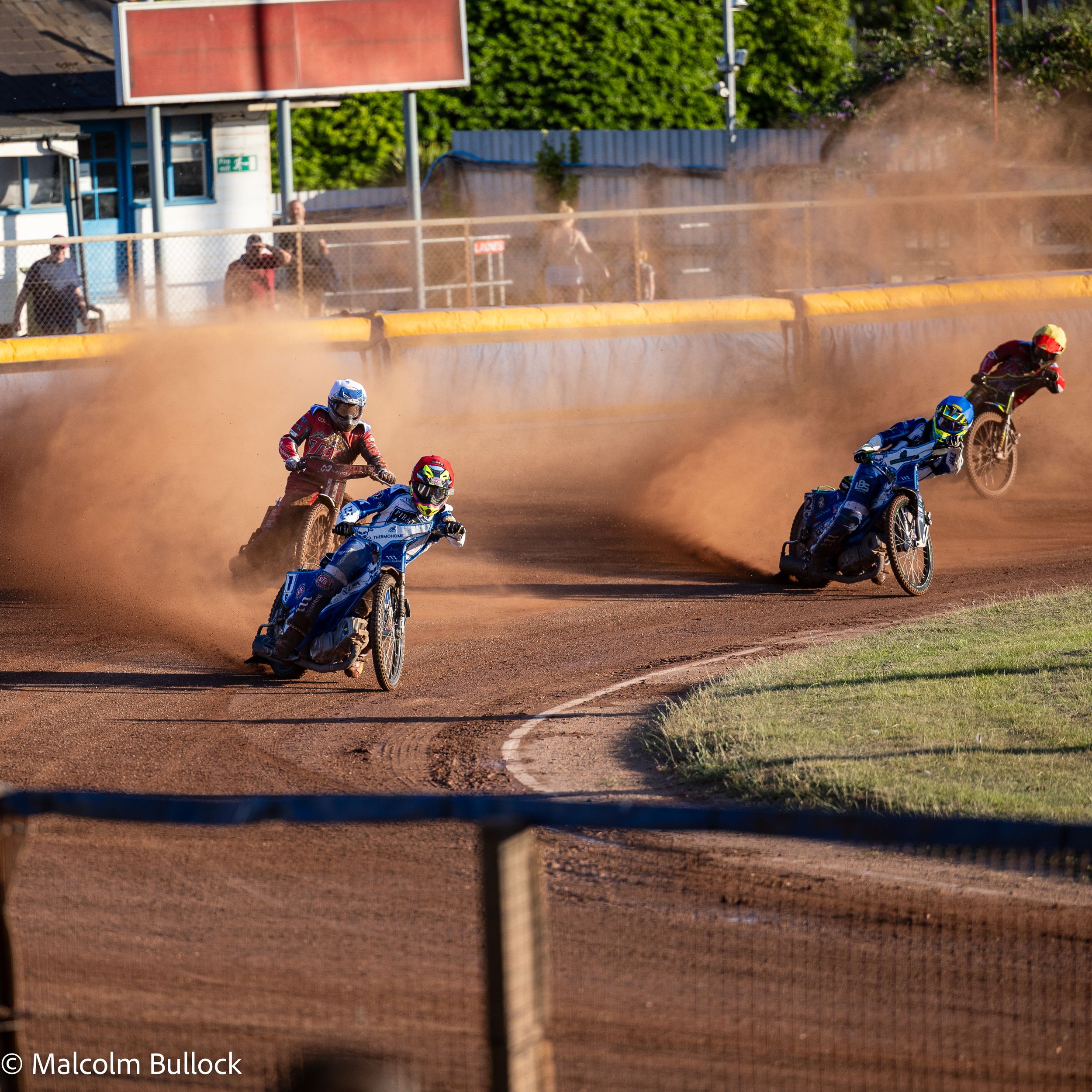 Three speedway riders on motorbikes kick up clouds of dust while racing on a dirt track at an evening motorsport event.