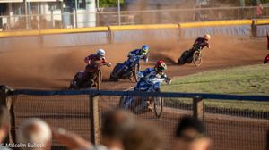 Multiple speedway motorcycle racers compete on a dirt oval track at dusk with spectators watching from the stands.
