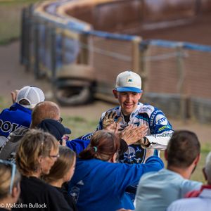 A smiling young speedway rider in a white cap high-fives fans over the barrier after a race at a dirt track venue.