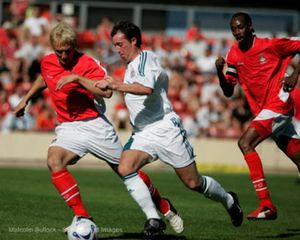Three soccer players compete for the ball during an intense match, with players in red and white uniforms on a sunny day.