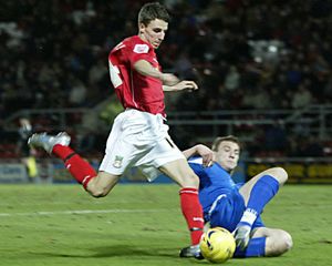 Soccer player in red kit dribbles past sliding tackle from opponent in blue during match.