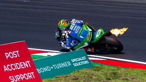 MotoGP rider on green motorcycle with flames shooting from exhaust on race track curve.