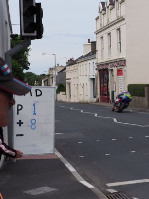 A motorcycle racer speeds through a narrow town street past a timing board showing position plus 8 during a road race.