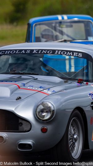 A classic silver Aston Martin DB4 race car sponsored by Nicholas King Homes leads a blue car on a racing circuit.