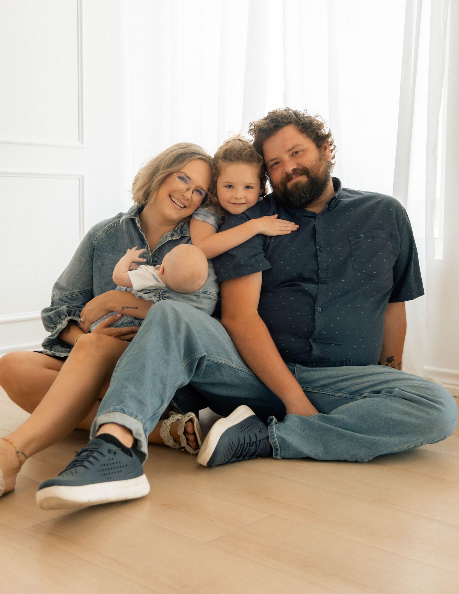 A happy family of four poses on a light wood floor, with parents and two young children smiling warmly in casual denim outfits.