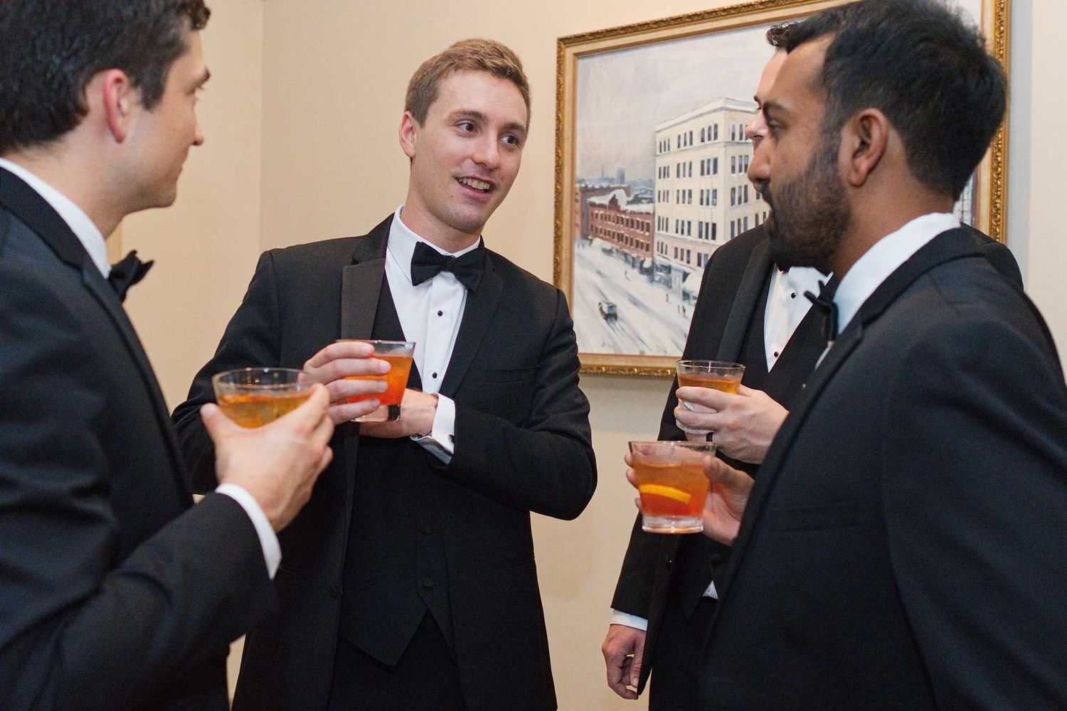 Four men in tuxedos socializing at a formal event, holding cocktails and conversing near a framed painting.