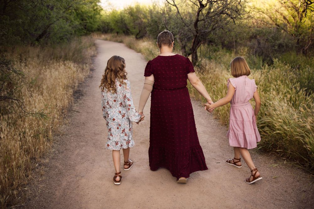 A woman in a burgundy maxi dress walks hand-in-hand with two girls in floral dresses along a scenic dirt path.