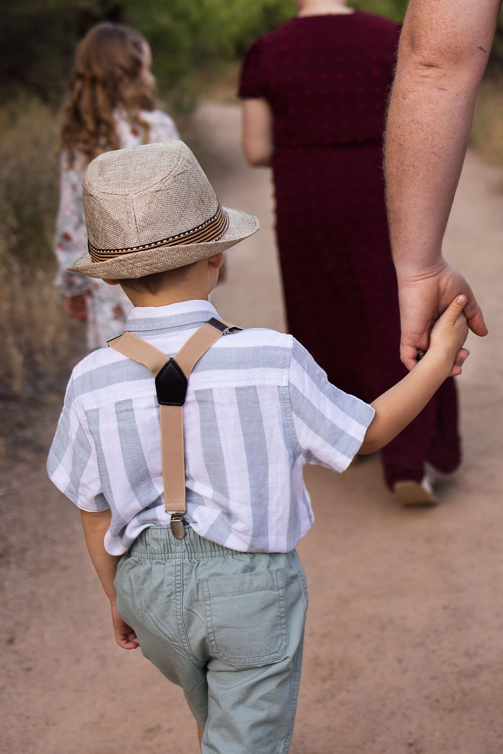 A young boy in a straw hat and suspenders holds an adults hand while walking on a desert trail.