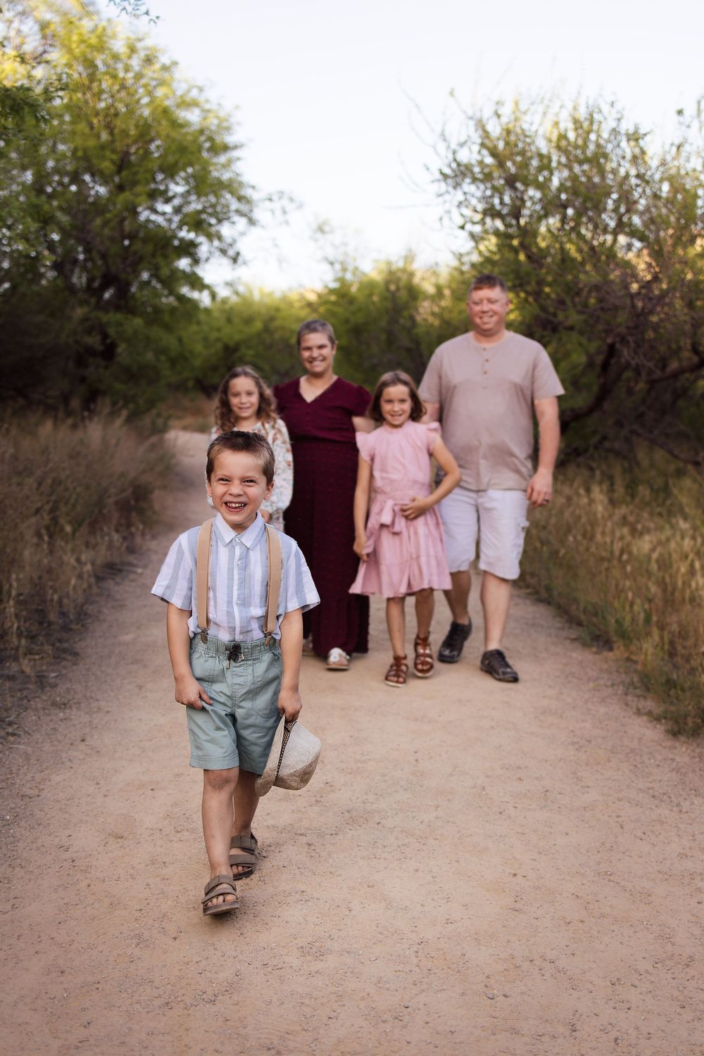 A joyful boy in suspenders leads his family down a scenic desert path surrounded by lush greenery.