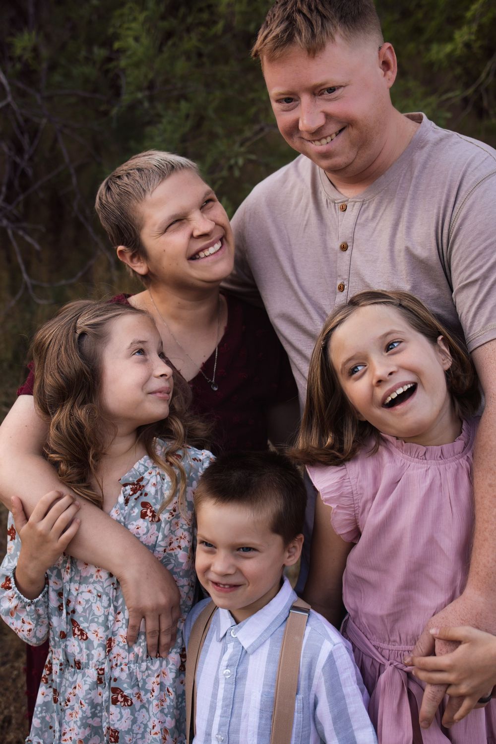 Happy family of five posing outdoors, two parents smiling with three young children in casual attire against a natural backdrop.