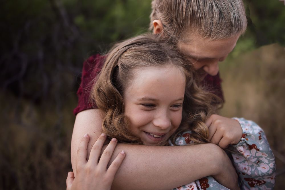 A young girl smiles joyfully while being hugged from behind by an older child outdoors.