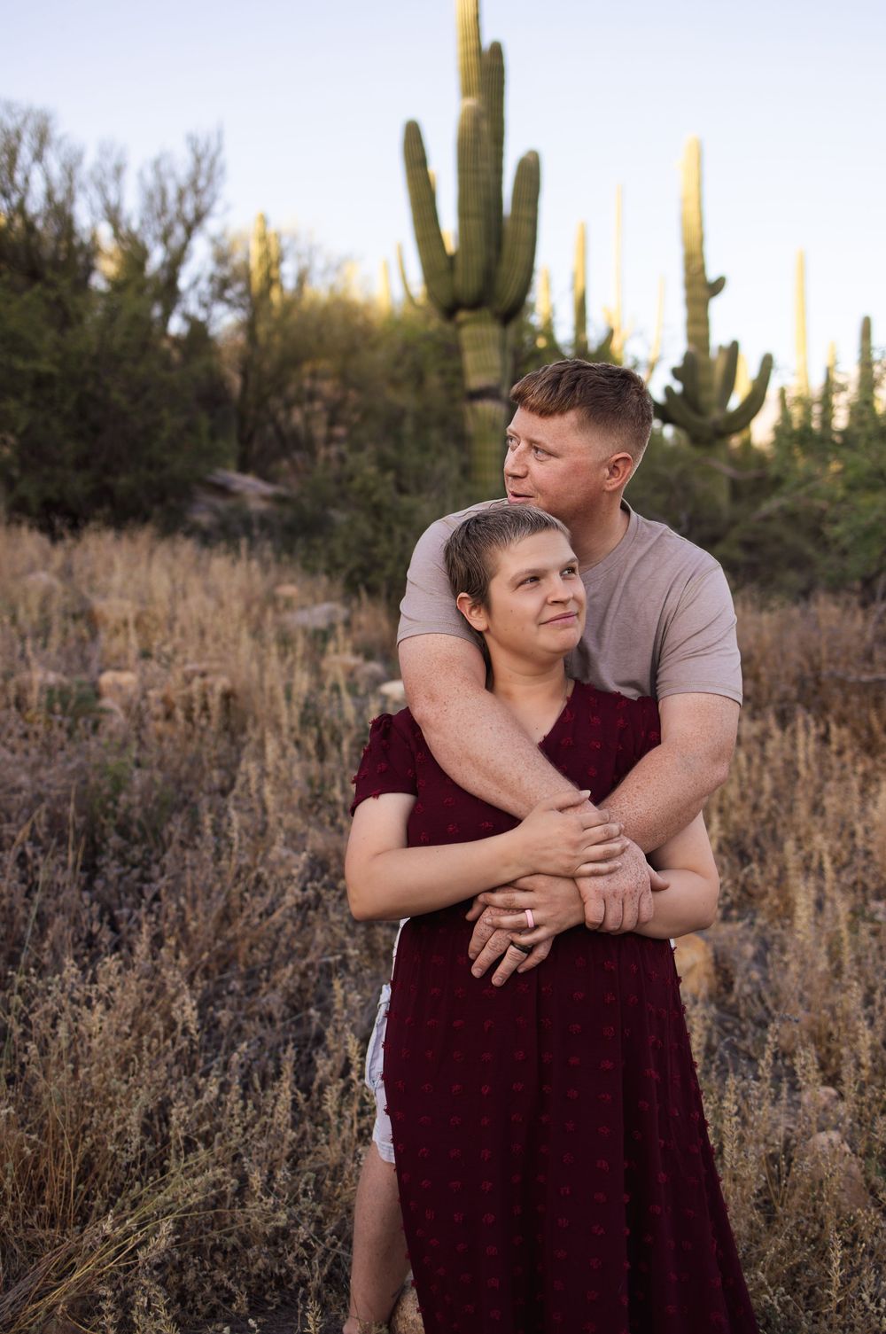 A couple embraces tenderly in a golden desert landscape with towering saguaro cacti at sunset.