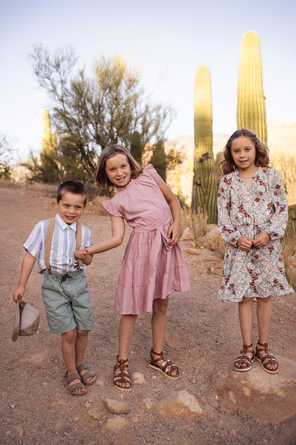 Three children pose in a desert landscape with tall saguaro cacti, dressed in pastel summer outfits and sandals.