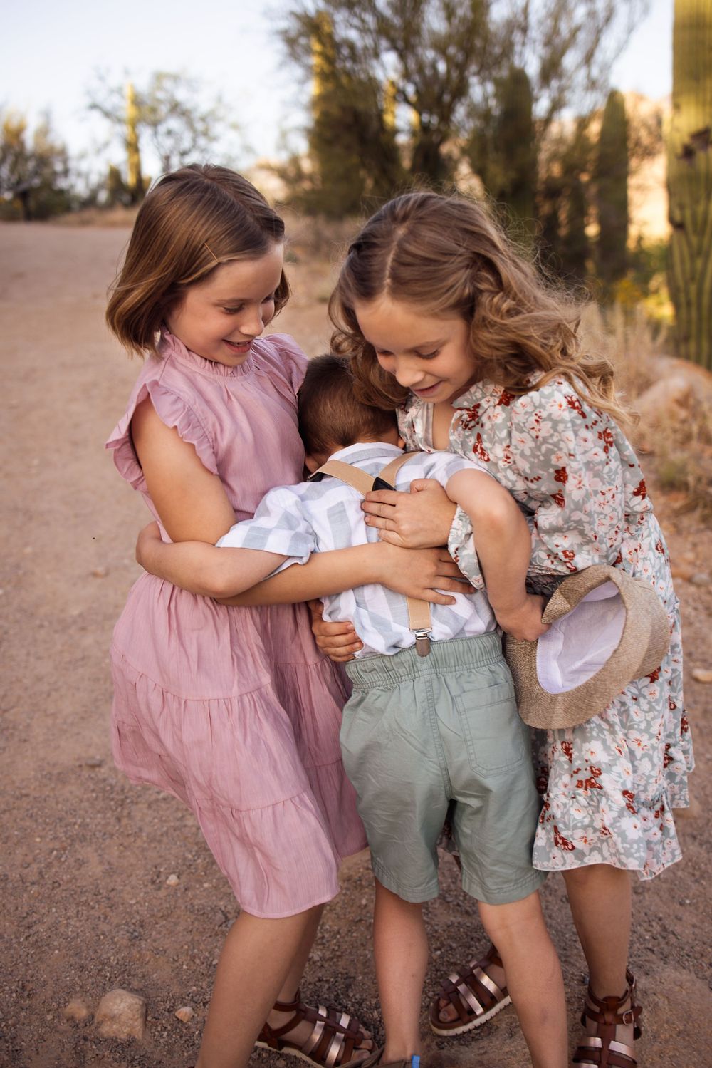 Three children share a warm group hug on a desert path, surrounded by saguaro cacti in soft golden light.