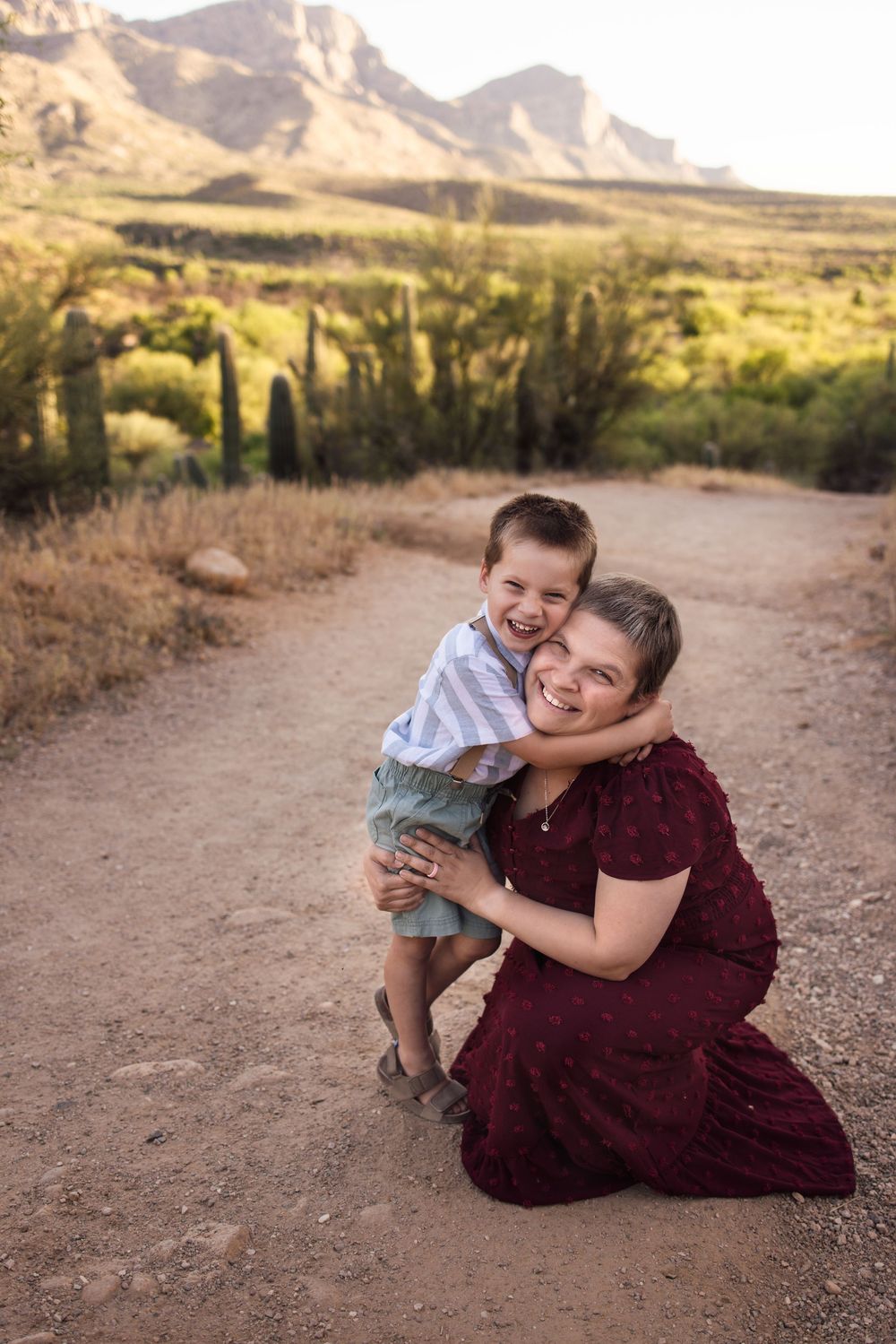 Woman in burgundy dress kneeling on desert path hugging laughing boy with saguaro cacti and mountains behind.