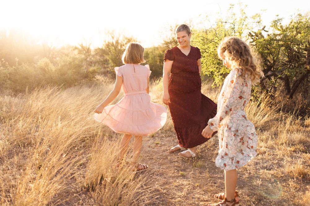 A woman in a black dress dances with two girls in a sunlit field during golden hour.