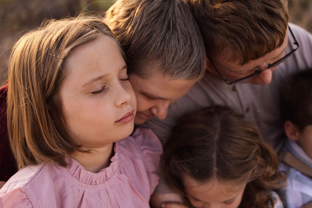 A father tenderly kisses his young daughter on the cheek during a warm family embrace outdoors.