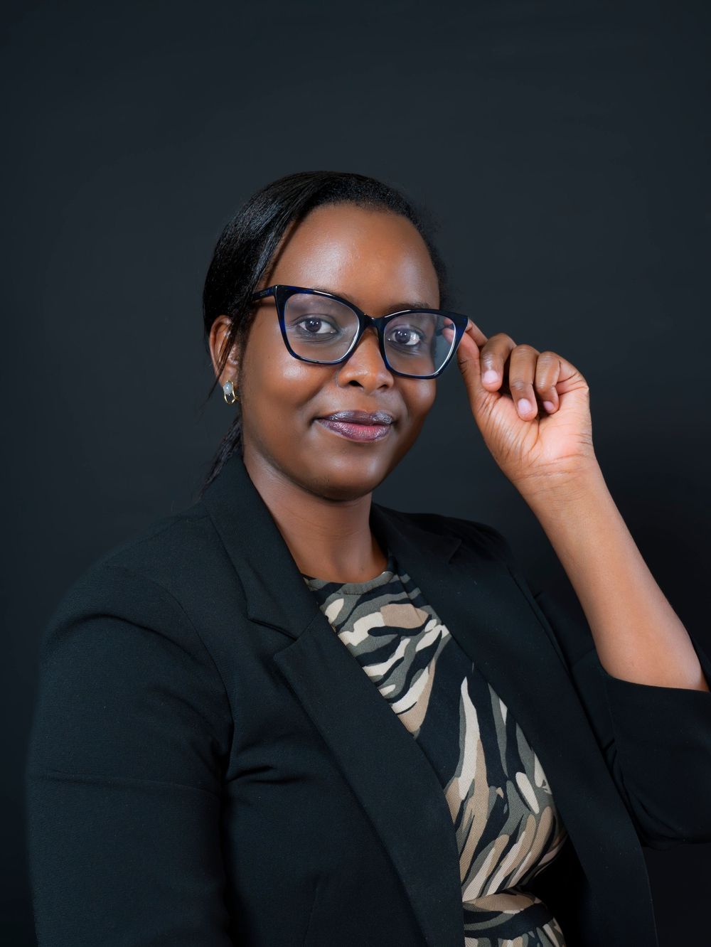 Professional African woman in black blazer adjusting glasses, posing confidently against dark background.