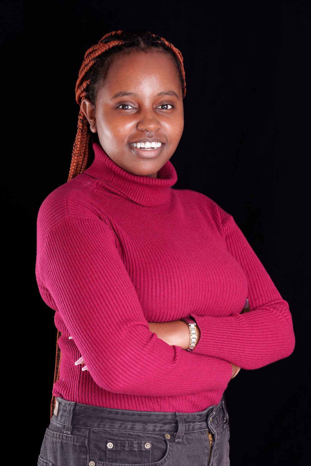 Young woman in pink turtleneck sweater with orange braids smiling with arms crossed.