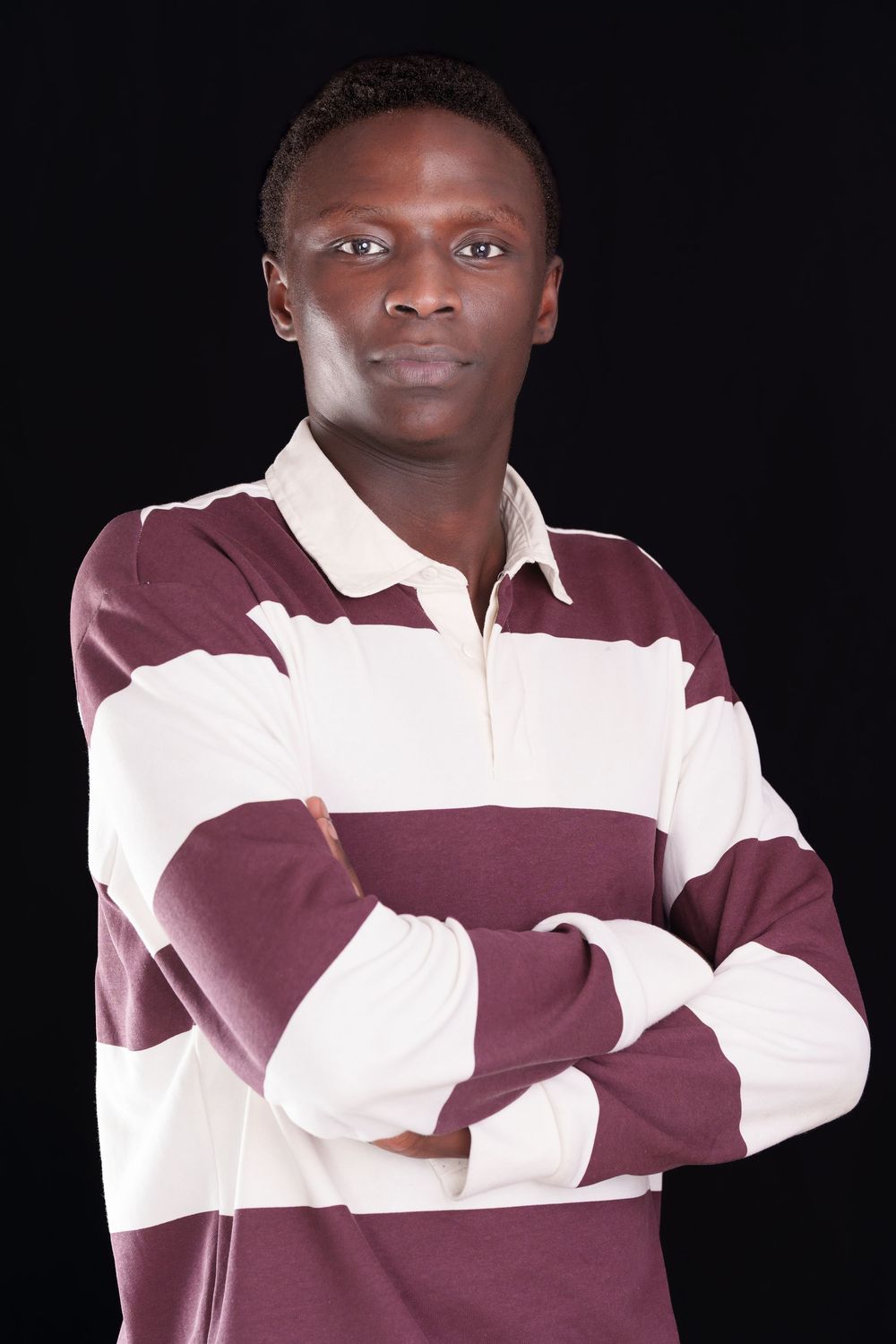 Young Black man in striped rugby shirt stands with arms crossed confidently against black background.
