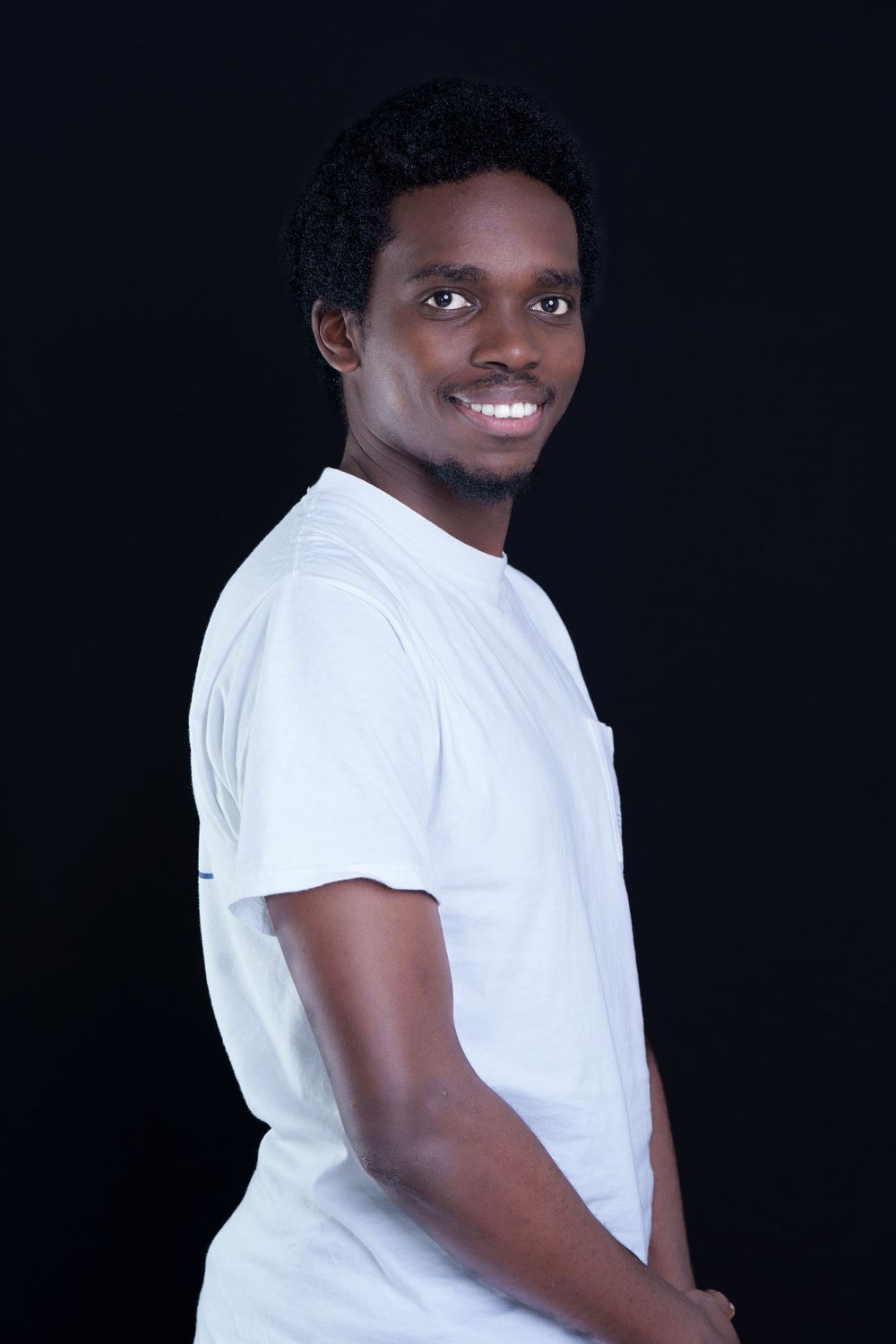 Smiling young Black man in white t-shirt poses in three-quarter turn against black background.