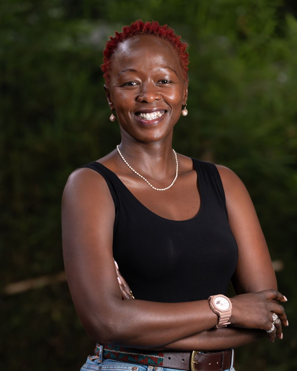Smiling Black woman with short red hair in black tank top poses outdoors with arms crossed.