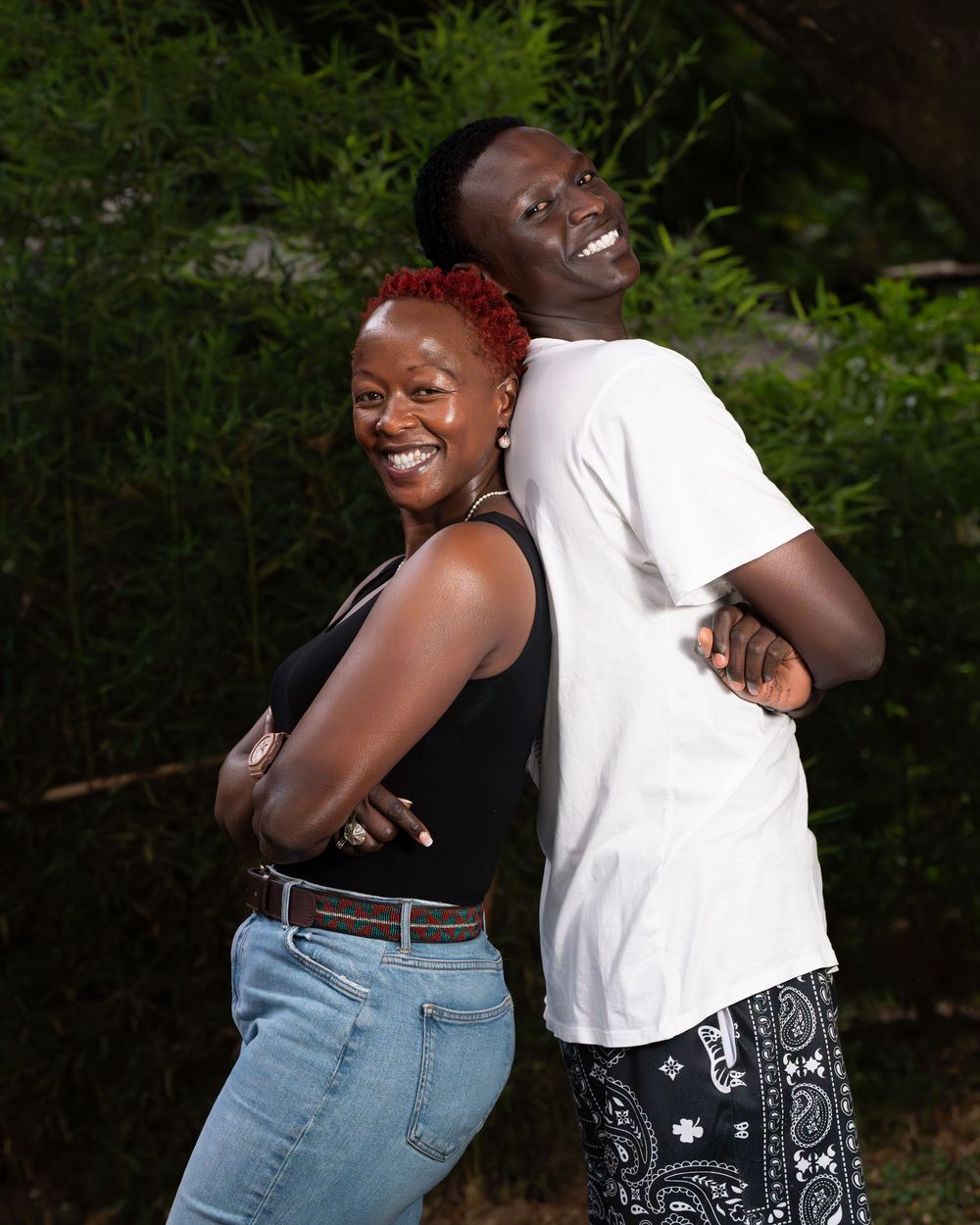Laughing Black woman and tall man pose back-to-back smiling outdoors in a lush green setting.