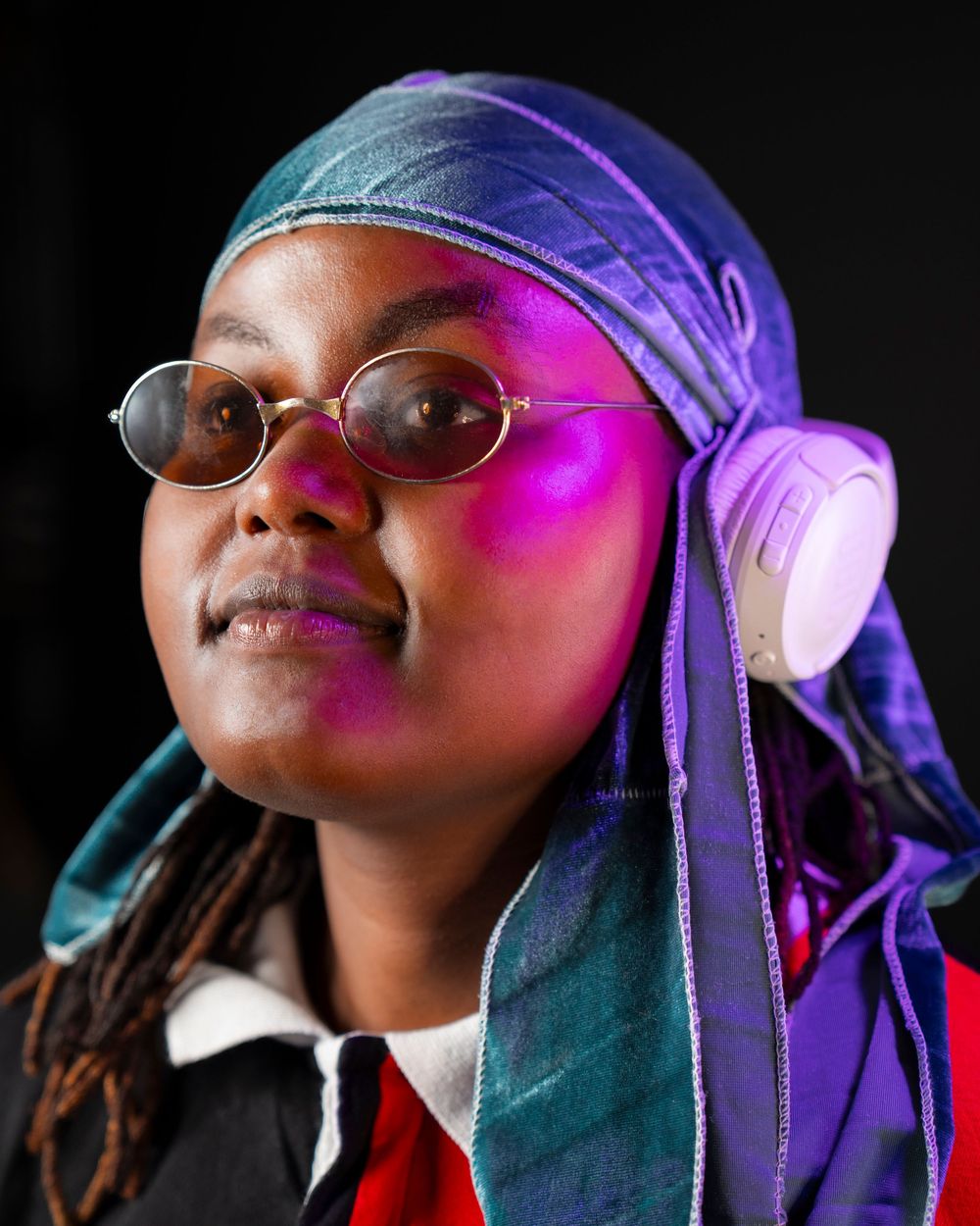 Black woman wearing colorful durag and round glasses illuminated by purple light with headphones around neck.
